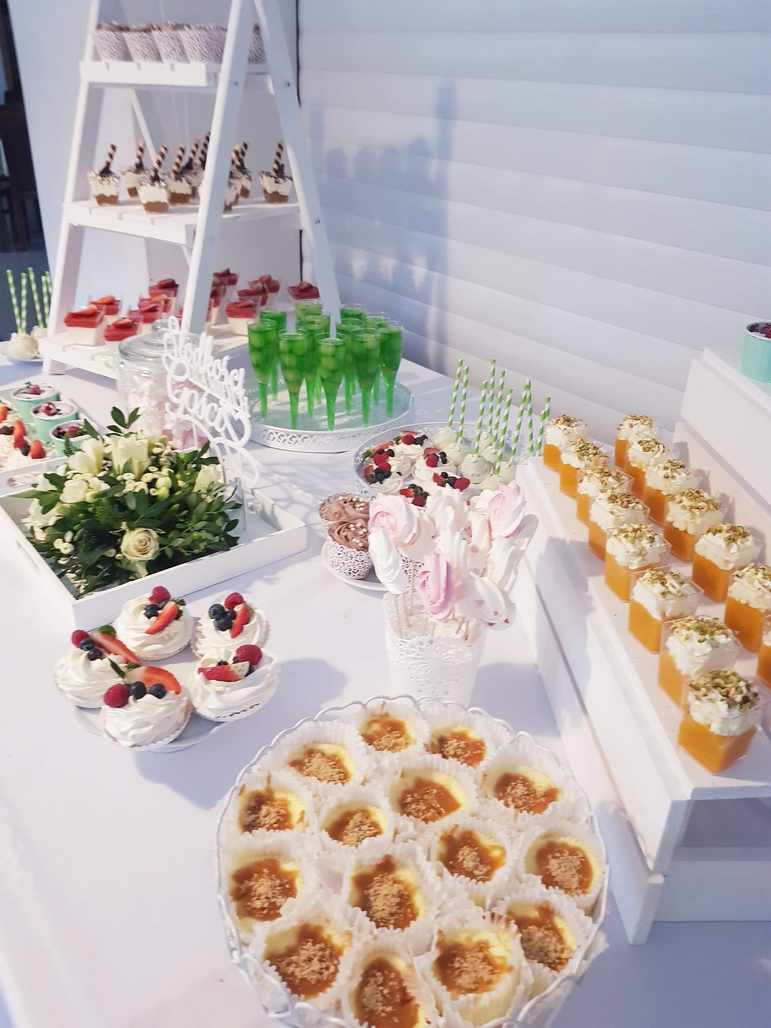 Słodki stół z różnorodnymi deserami: mini bezy z owocami, puddingi w kieliszkach, lizaki bezowe, mini serniczki z karmelem, zielone napoje w stożkowych szklankach, ułożone na białych podestach...