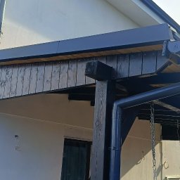 matdach.pl - Nowoczesna konstrukcja z ciemnego drewna, z widocznymi rynnami i elementami obróbki blacharskiej, przylegająca do białego budynku z oknem.