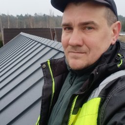 matdach.pl - Pracownik w odblaskowej kurtce i czapce z logo Velux na tle szarego dachu z blachy na rąbek stojący, w tle drzewa i pochmurne niebo.