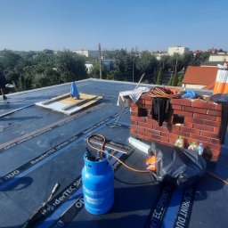 matdach.pl - Prace dekarskie na płaskim dachu pokrytym czarną papą BauderTEC Sprint Duo, widoczne butle gazowe, narzędzia, ceglany komin i dwóch dekarzy w oddali, w tle zabudowa miejska