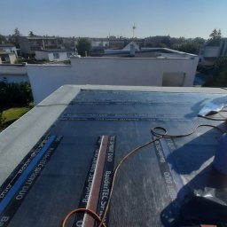 matdach.pl - Prace na płaskim dachu: aplikacja papy termozgrzewalnej BauderTEC Sprint DUO, widoczny palnik dekarski, butla gazowa i częściowo odsłonięta sylwetka dekarza na tle zabudowy miejskiej.