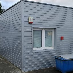 matdach.pl - Szary, prefabrykowany budynek z blachy trapezowej z oknem, alarmem pożarowym i drzwiami wejściowymi, widoczny kontener na śmieci i samochód dostawczy w tle.