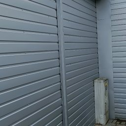 matdach.pl - Szara, blaszana elewacja budynku z widocznym ujściem wentylacji dachowej, rynną i fragmentem chodnika z kostki brukowej.