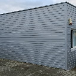 matdach.pl - Szara, blaszana konstrukcja budynku z widoczną ścianą z poziomych paneli blaszanych, oknem i dwoma wejściami, na zewnątrz widoczny kosz na śmieci i butle z gazem w metalowej klatce.