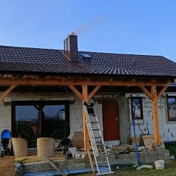 matdach.pl - Dom w trakcie budowy z nowym, brązowym dachem z blachodachówki, drewnianą konstrukcją werandy i kominem, z którego wydobywa się dym.