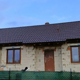 matdach.pl - Dom w trakcie budowy z brązowym dachem z blachodachówki, widoczne okna z roletami zewnętrznymi i fragment elewacji z białego bloczka, w tle pochmurne niebo.