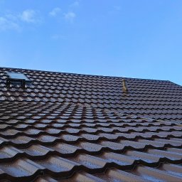 matdach.pl - Nowy, brązowy dach z blachodachówki z kominem, wyłazem dachowym i odpowietrznikiem, widoczne krople wody na powierzchni, w tle niebieskie niebo i fragment dachu sąsiedniego budynku zabezpieczonego...