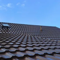 Nowy, brązowy dach z blachodachówki z kominem, wyłazem dachowym i odpowietrznikiem, widoczne krople wody na powierzchni, w tle niebieskie niebo i fragment dachu sąsiedniego budynku zabezpieczonego...