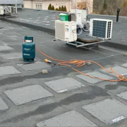 Bitumiczny dach płaski z widocznymi łatami, butlą gazową i palnikiem dekarskim podczas prac, w tle klimatyzatory na wspornikach.