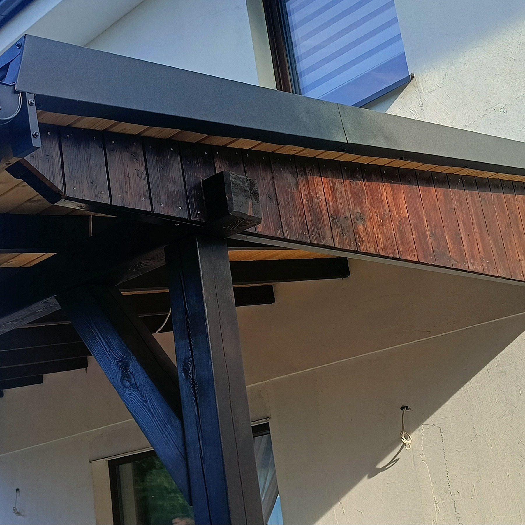 Drewniana konstrukcja z elementami obróbki blacharskiej, widok od dołu na zadaszenie tarasu z ciemnego drewna i blachy w kolorze antracytowym, widoczne mocowania i elementy konstrukcyjne.