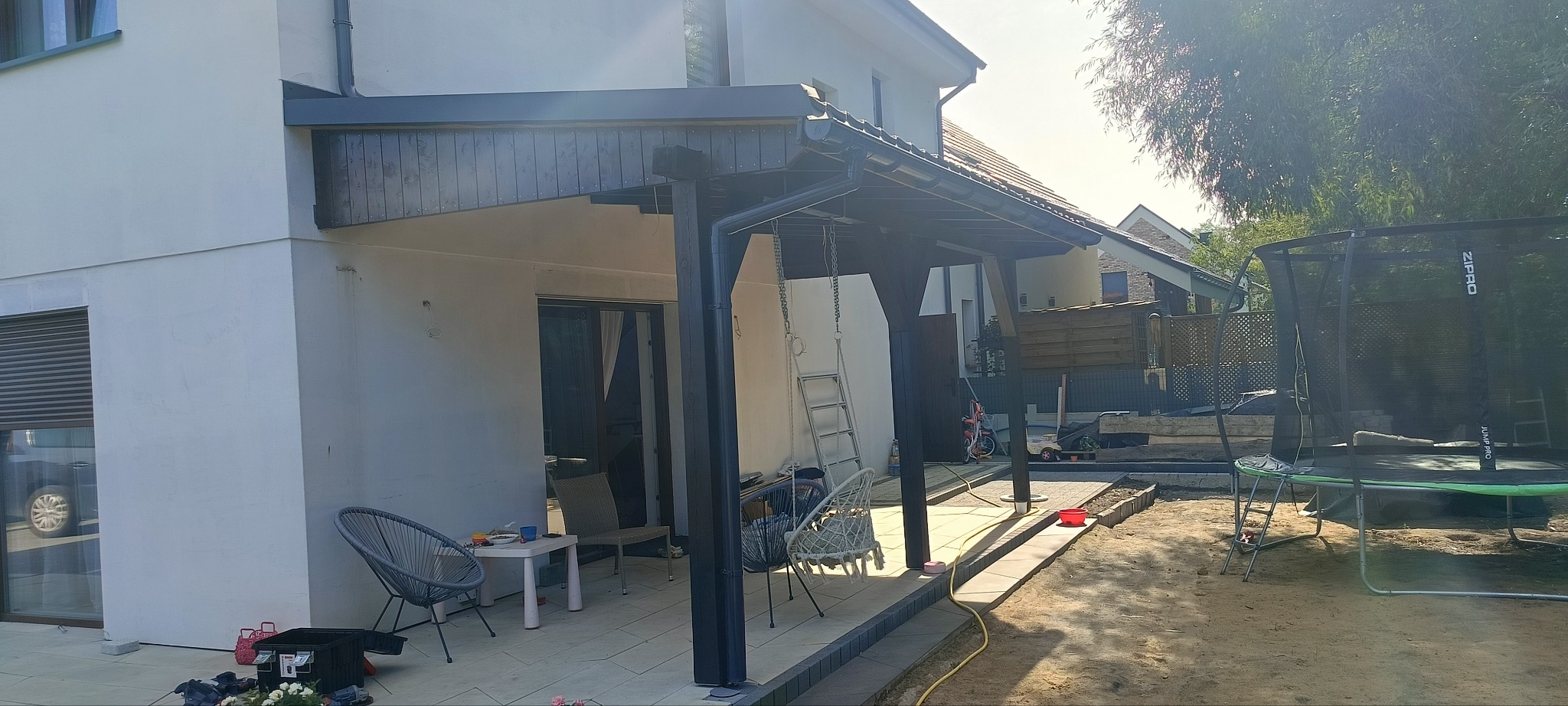 Zadaszenie tarasu z ciemnego drewna, rynna w kolorze grafitowym, biała elewacja budynku, widoczne elementy ogrodu: trampolina i meble ogrodowe.