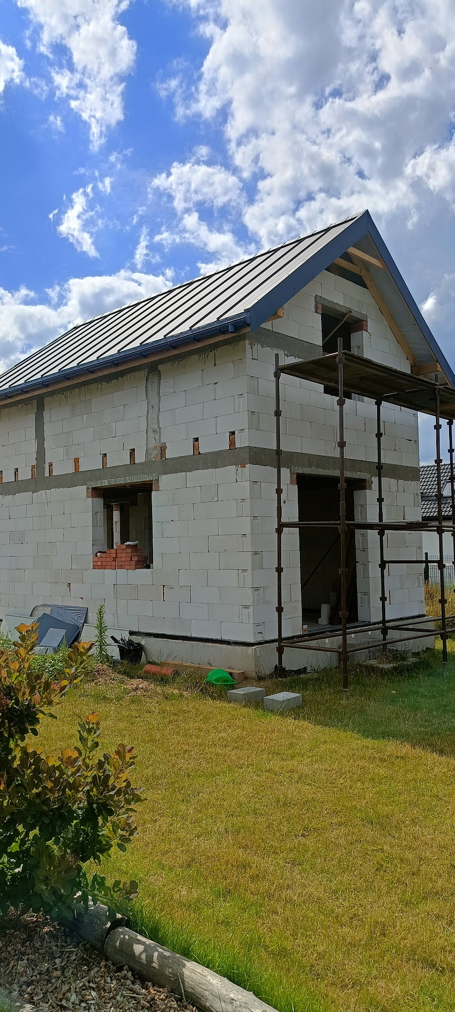 Budowa domu z białych bloczków, widoczny szkielet dachu pokryty czarną blachą, rusztowanie, nieotynkowane ściany, trawa i niebo z chmurami.