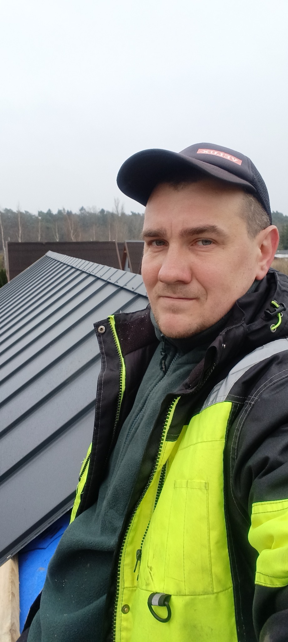 Pracownik w odblaskowej kurtce i czapce z logo Velux na tle szarego dachu z blachy na rąbek stojący, w tle drzewa i pochmurne niebo.