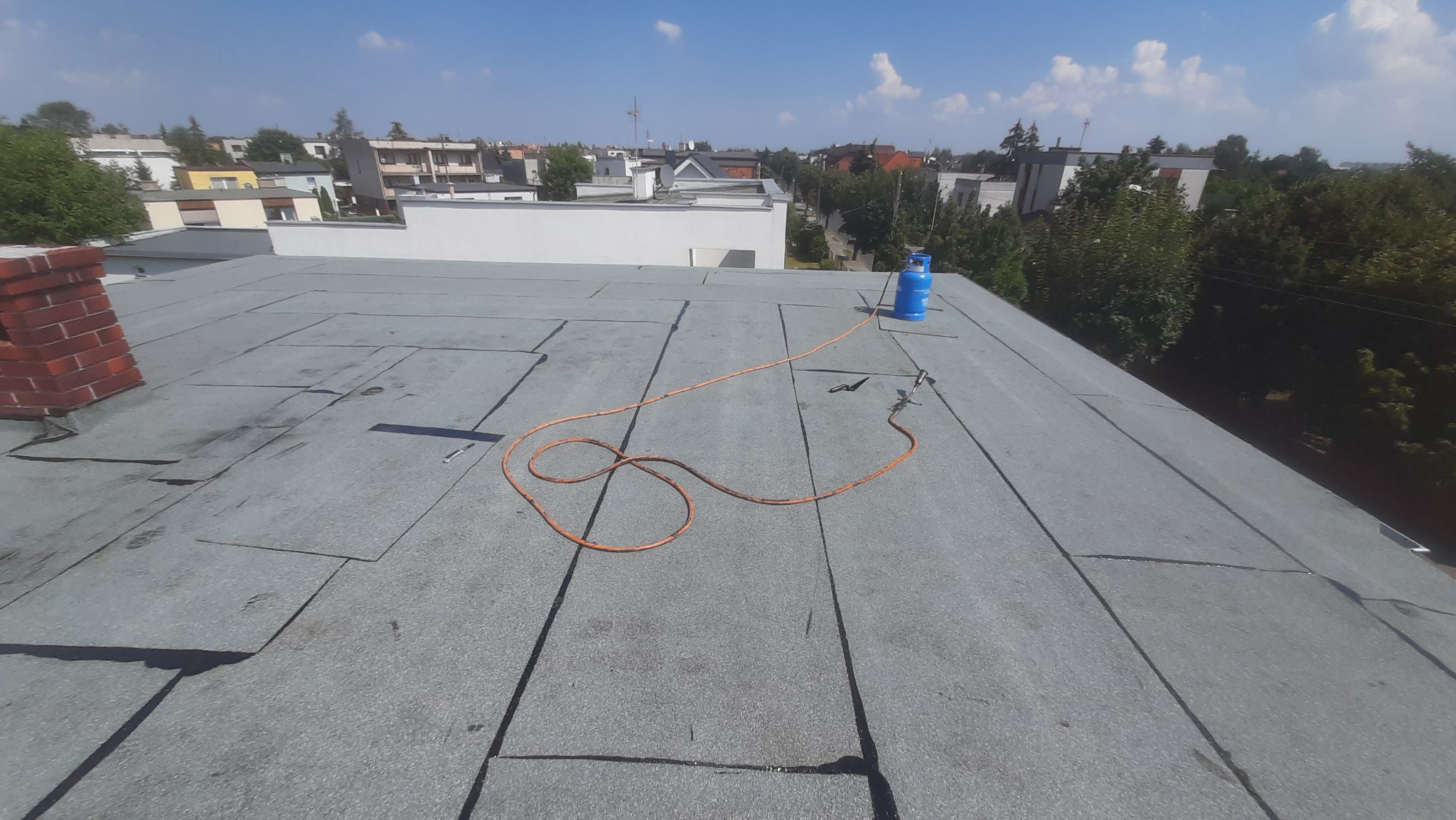 Płaski dach pokryty papą termozgrzewalną z widocznym palnikiem dekarskim podłączonym do niebieskiej butli gazowej, komin z czerwonej cegły w tle.