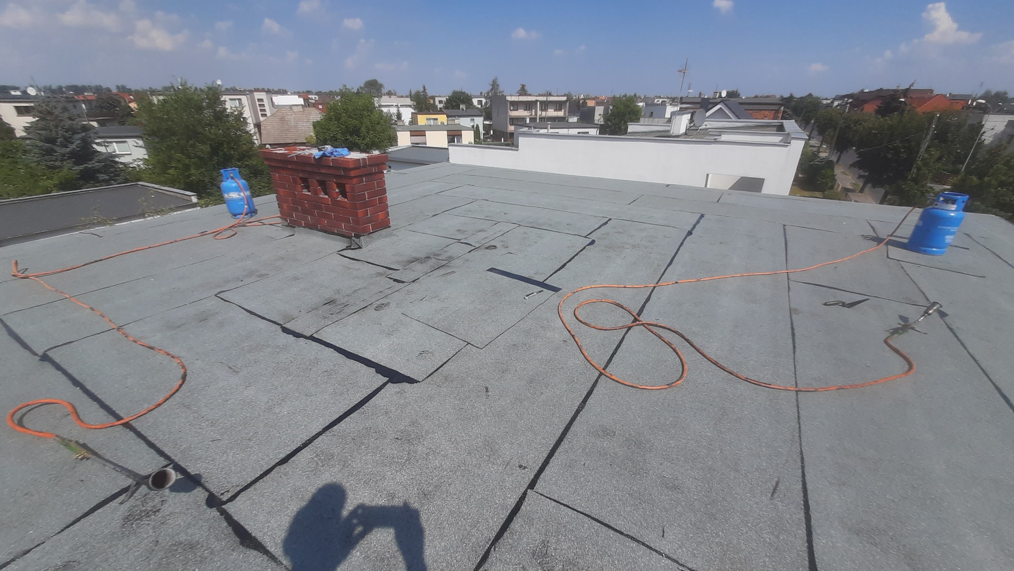 Płaski dach pokryty papą termozgrzewalną z widocznym kominem, butlami gazowymi i narzędziami do zgrzewania papy, w tle zabudowa miejska.