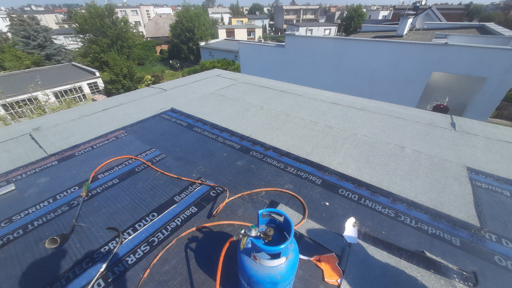 Prace na płaskim dachu: membrana bitumiczna BauderTEC SPRINT DUO, palnik dekarski podłączony do niebieskiej butli gazowej, widok z góry na tle zabudowy miejskiej.