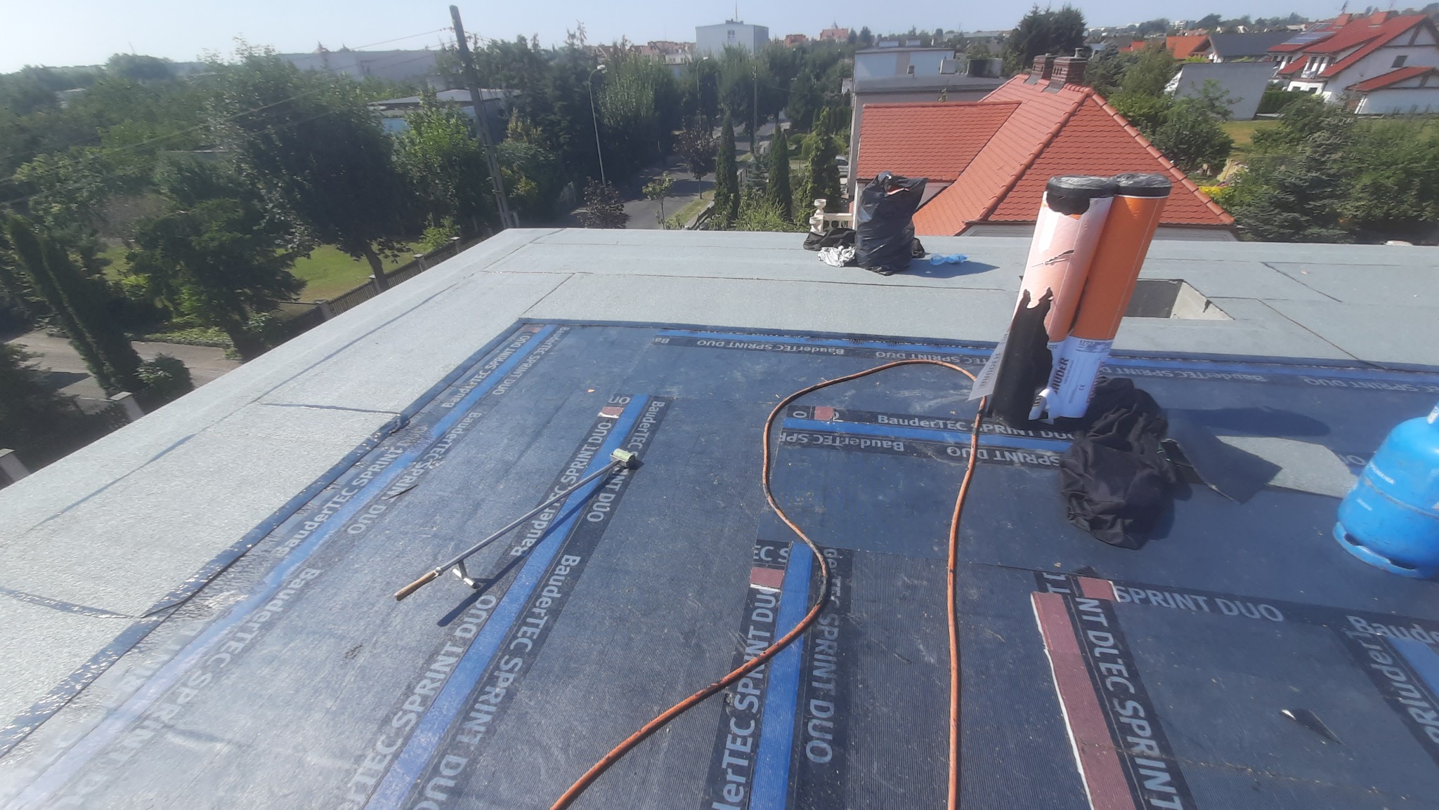 Płaski dach pokryty papą termozgrzewalną BauderTEC Sprint DUO, widoczne narzędzia dekarskie, butla gazowa i rolki papy na tle zabudowy miejskiej.