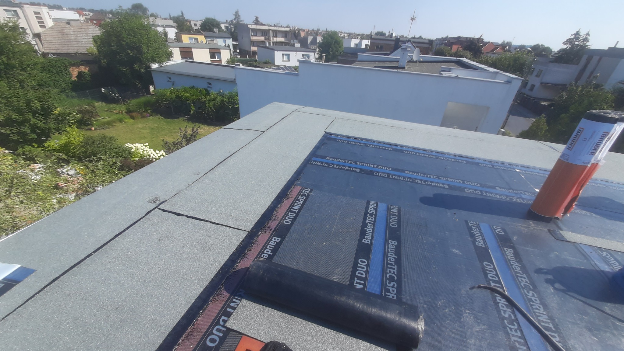 Montaż papy termozgrzewalnej BauderTEC Sprint Duo na płaskim dachu, widok z góry na częściowo pokrytą powierzchnię z rolką papy i narzędziami w tle zabudowa miejska.