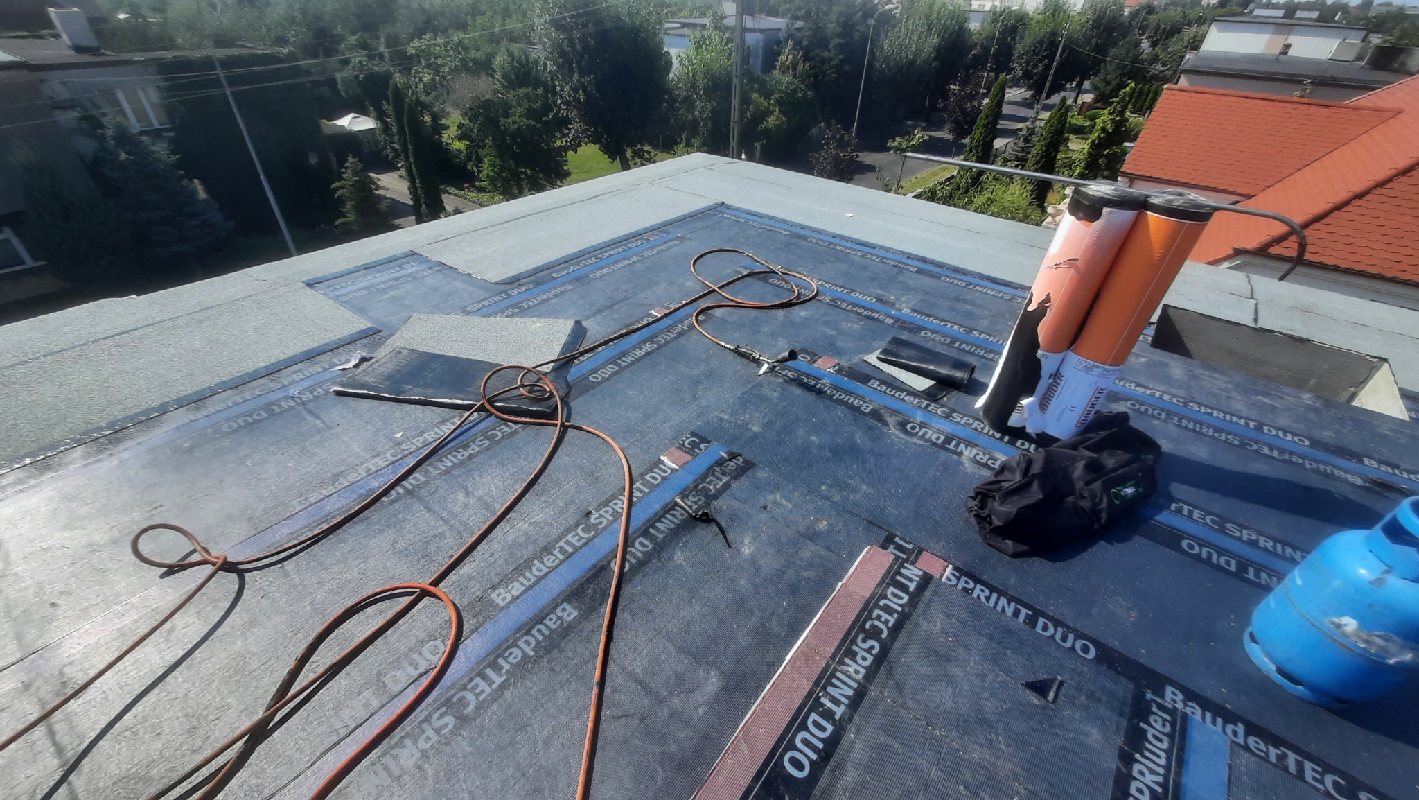 Płaski dach pokryty czarną papą termozgrzewalną z widocznymi zgrzewami, leżące obok rolki papy, butla gazowa i przewód palnika dekarskiego, w tle zabudowa miejska i zieleń.