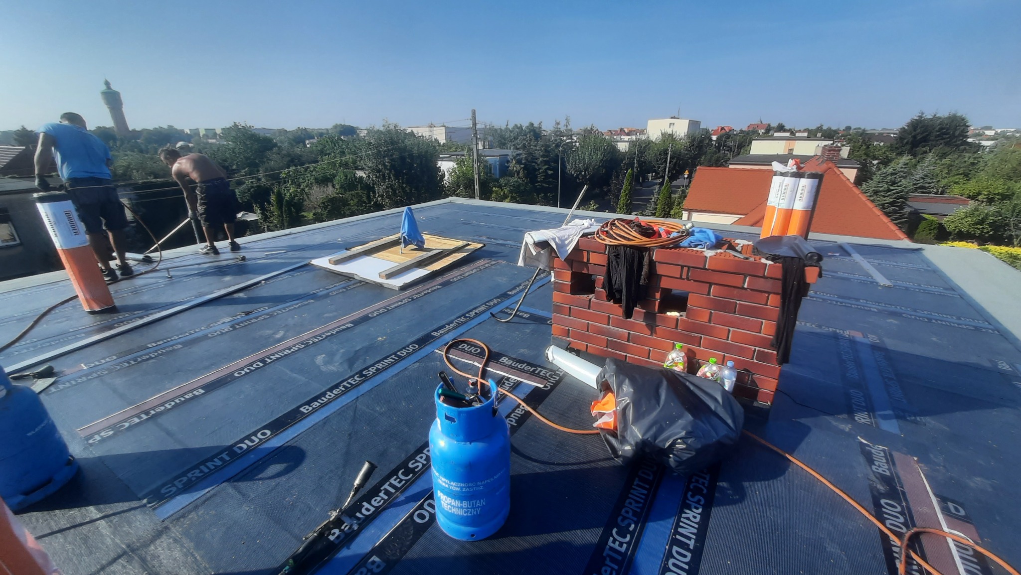 Prace dekarskie na płaskim dachu pokrytym czarną papą BauderTEC Sprint Duo, widoczne butle gazowe, narzędzia, ceglany komin i dwóch dekarzy w oddali, w tle zabudowa miejska