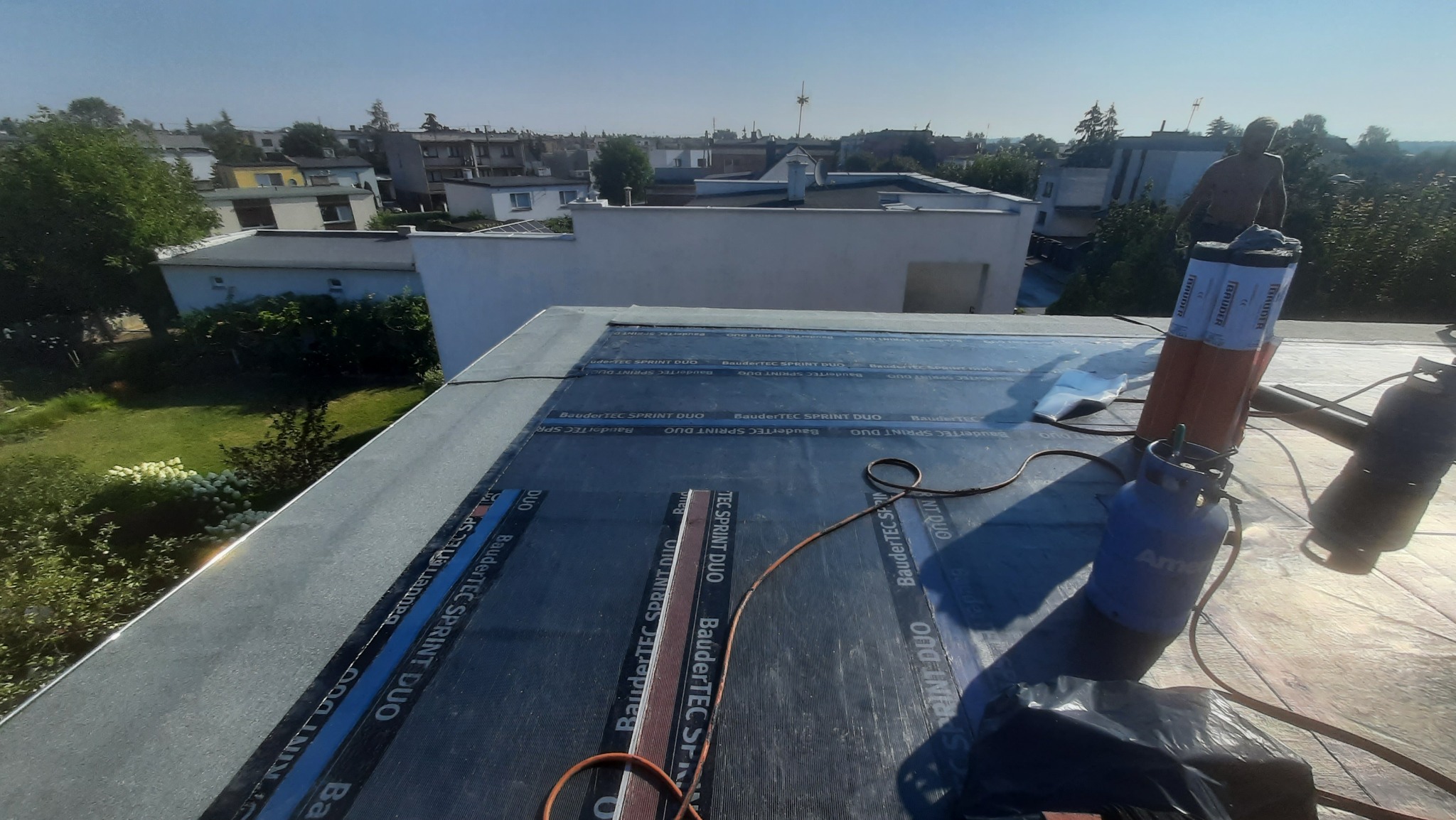 Prace na płaskim dachu: aplikacja papy termozgrzewalnej BauderTEC Sprint DUO, widoczny palnik dekarski, butla gazowa i częściowo odsłonięta sylwetka dekarza na tle zabudowy miejskiej.