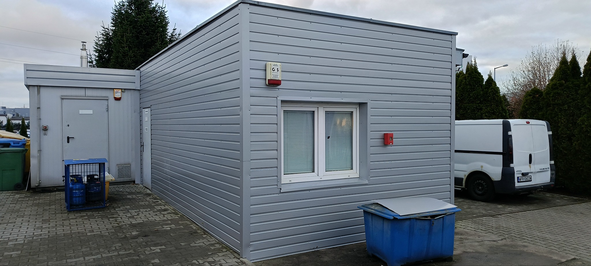 Szary, prefabrykowany budynek z blachy trapezowej z oknem, alarmem pożarowym i drzwiami wejściowymi, widoczny kontener na śmieci i samochód dostawczy w tle.