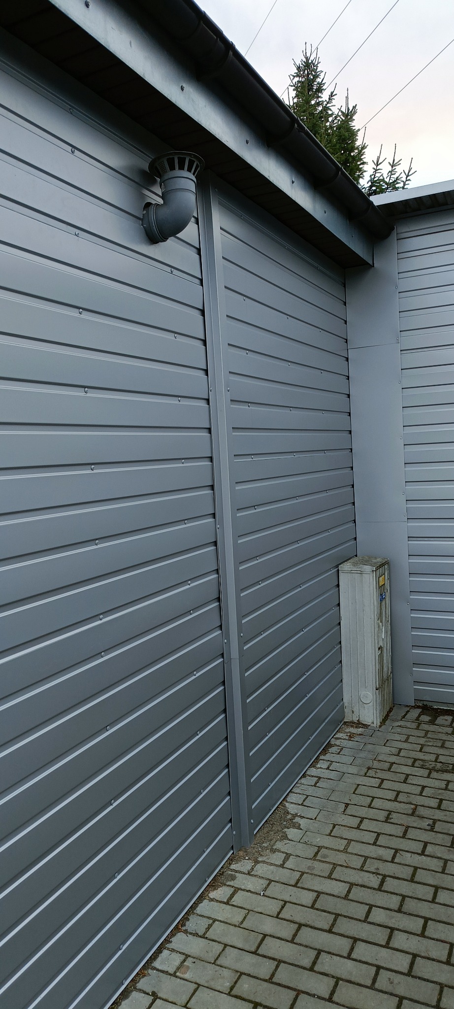 Szara, blaszana elewacja budynku z widocznym ujściem wentylacji dachowej, rynną i fragmentem chodnika z kostki brukowej.