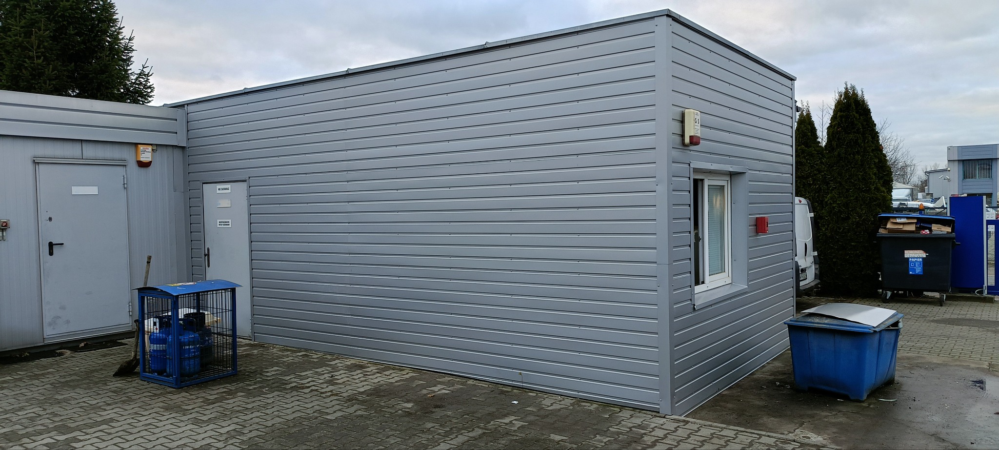Szara, blaszana konstrukcja budynku z widoczną ścianą z poziomych paneli blaszanych, oknem i dwoma wejściami, na zewnątrz widoczny kosz na śmieci i butle z gazem w metalowej klatce.