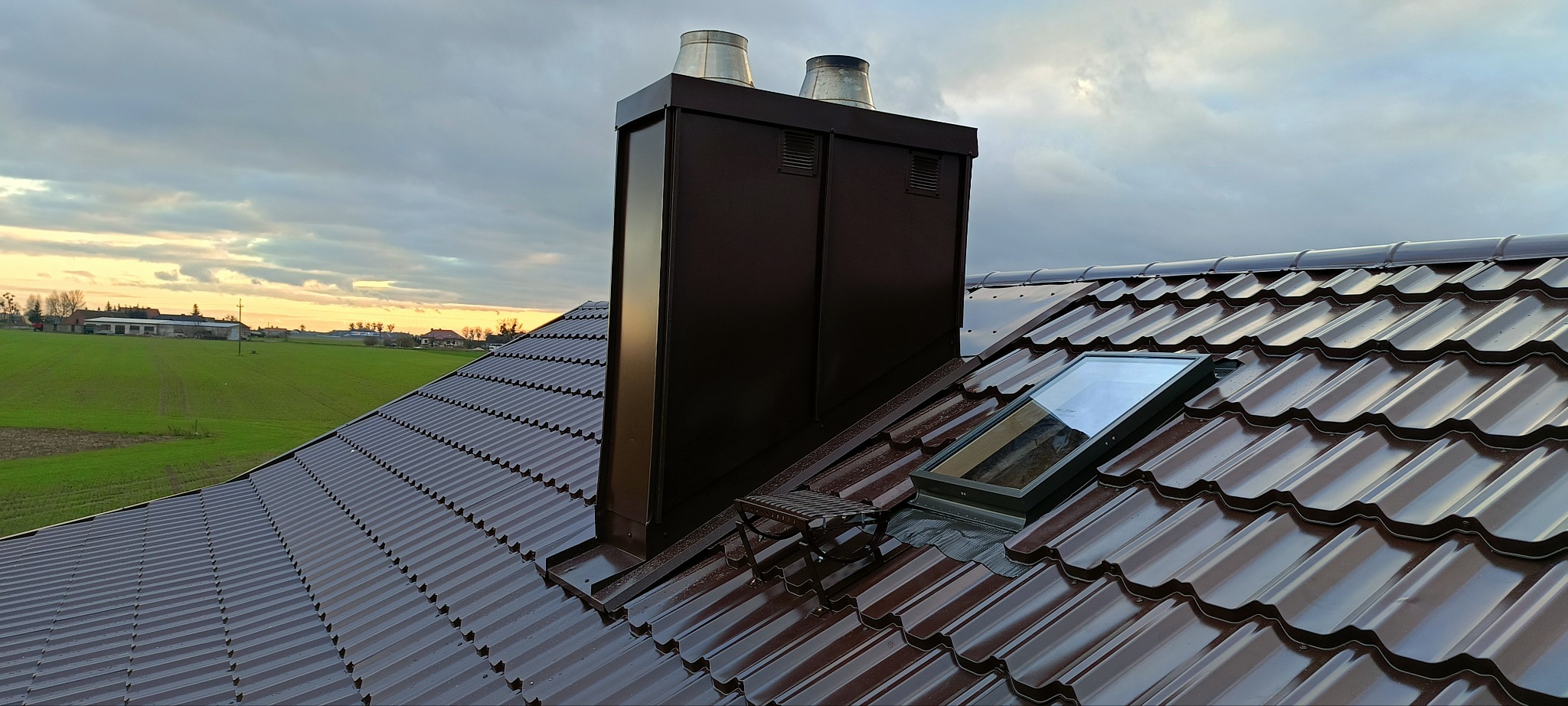 Brązowy dach z blachy trapezowej, komin z dwoma wylotami, okno dachowe z widocznym niebem, krajobraz wiejski w tle pod pochmurnym niebem.