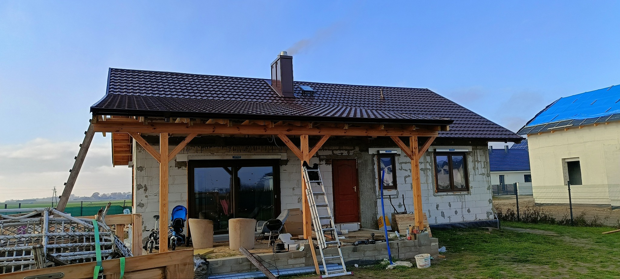 Dom w trakcie budowy z nowym, brązowym dachem z blachodachówki, drewnianą konstrukcją werandy i kominem, z którego wydobywa się dym.