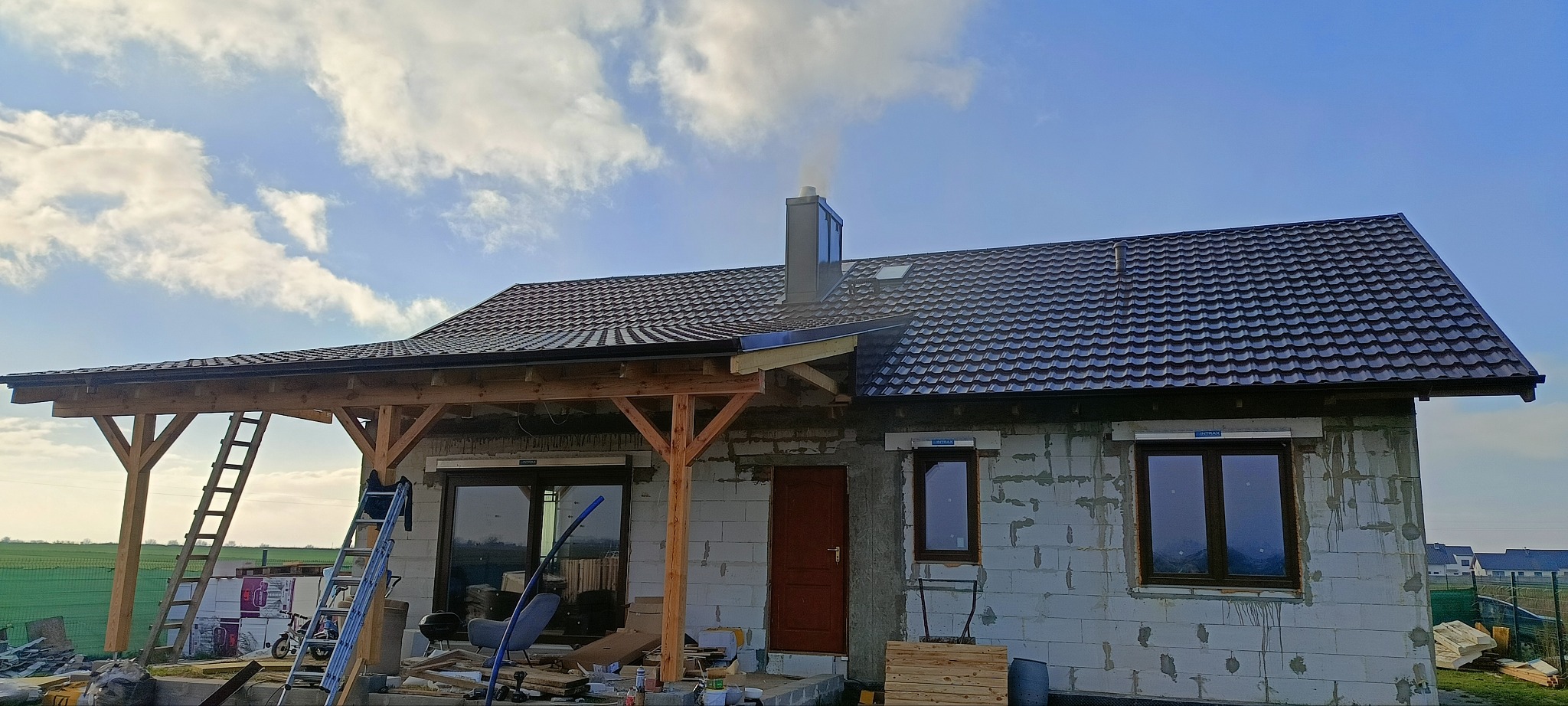 Dom w trakcie krycia dachu dachówką modułową w kolorze grafitowym, widoczny komin z dymem, rusztowanie, elementy konstrukcji zadaszenia tarasu i materiały budowlane.