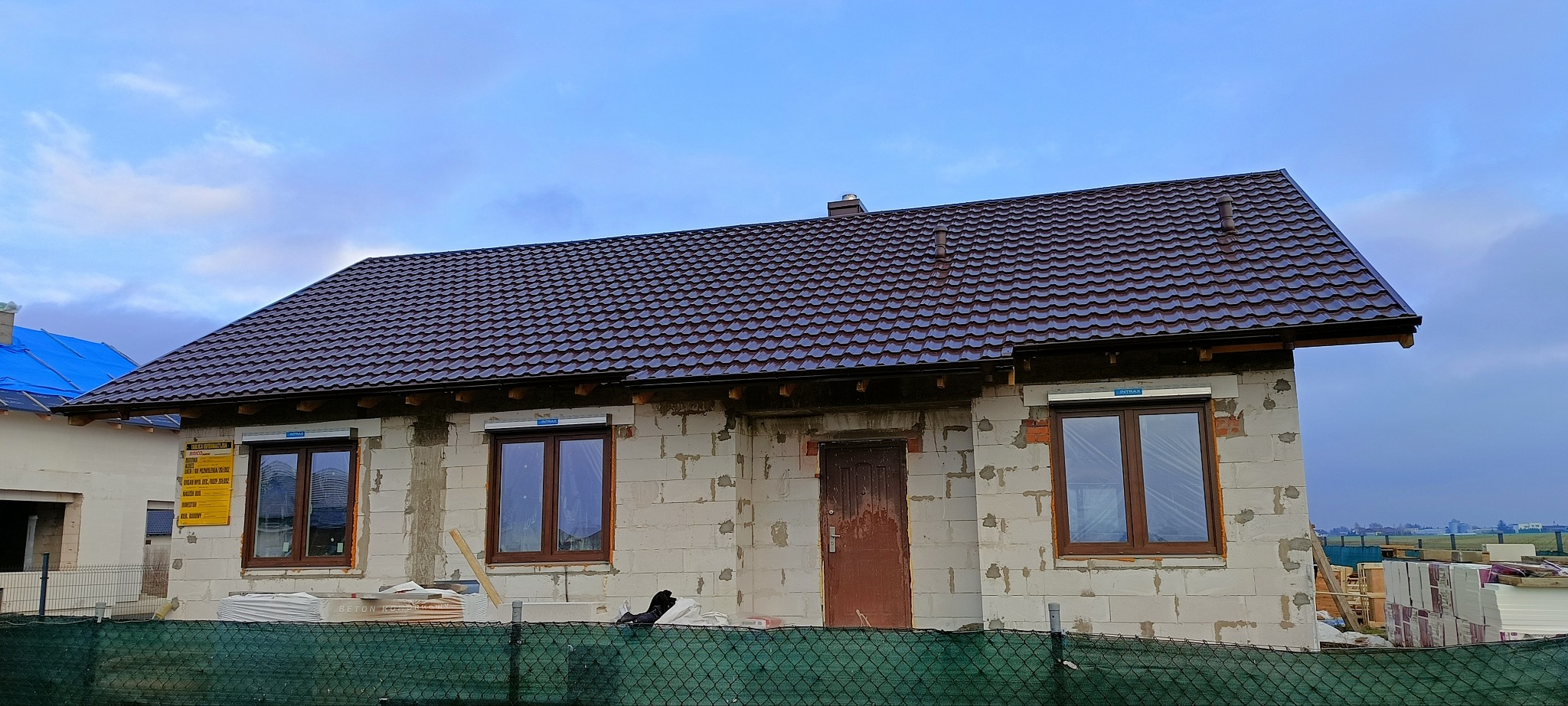 Dom w trakcie budowy z brązowym dachem z blachodachówki, widoczne okna z roletami zewnętrznymi i fragment elewacji z białego bloczka, w tle pochmurne niebo.