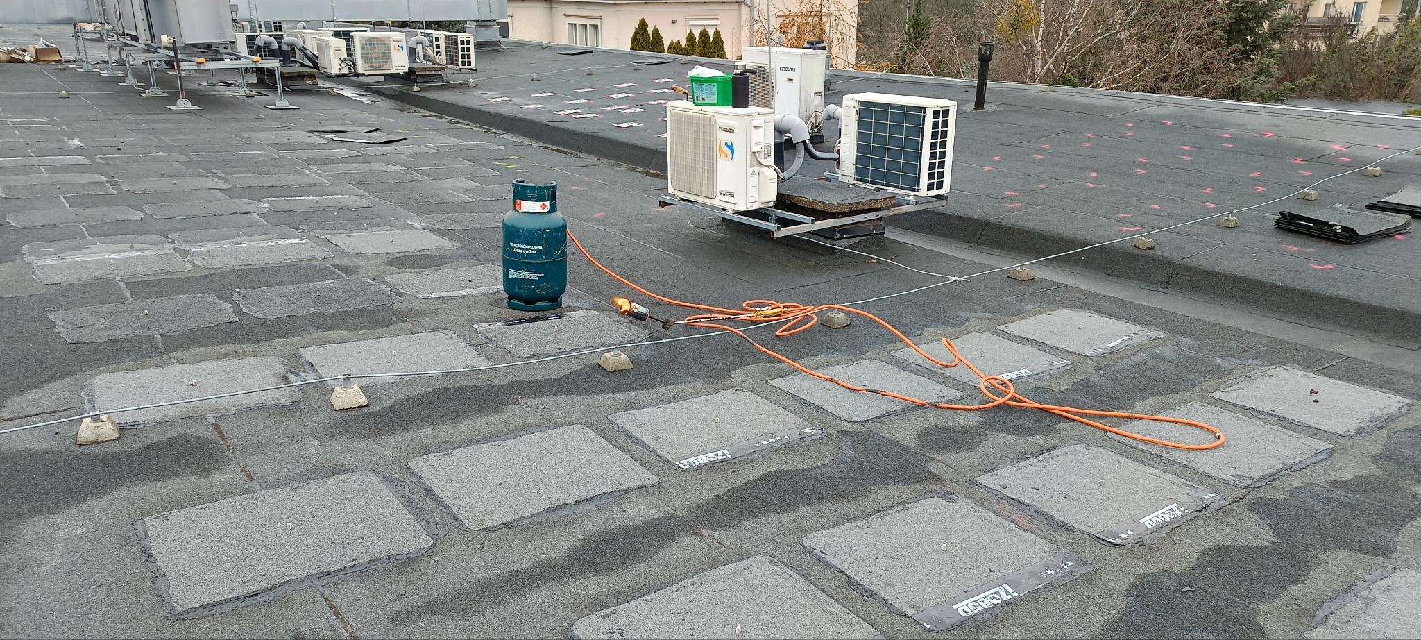 Bitumiczny dach płaski z widocznymi łatami, butlą gazową i palnikiem dekarskim podczas prac, w tle klimatyzatory na wspornikach.