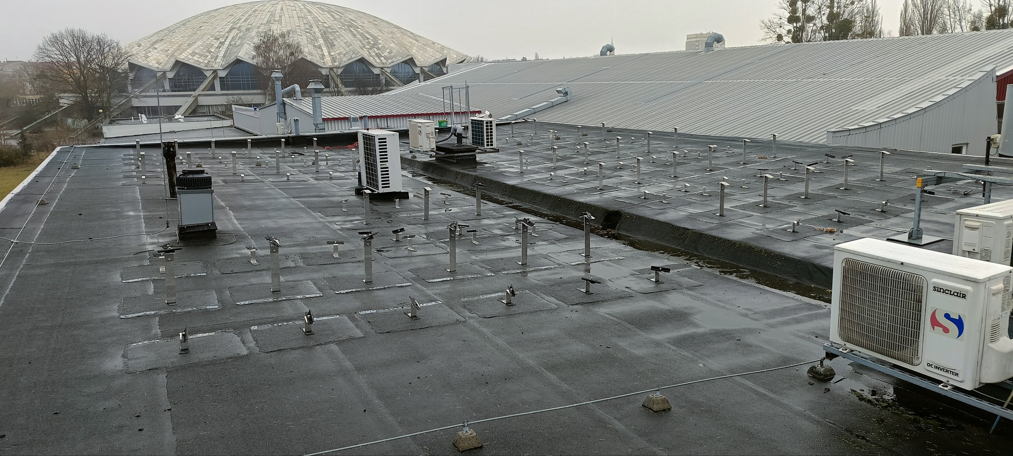 Płaski dach pokryty papą z licznymi stalowymi wspornikami, wentylatorami i klimatyzatorami Sinclair. W tle widoczny budynek z kopułą.