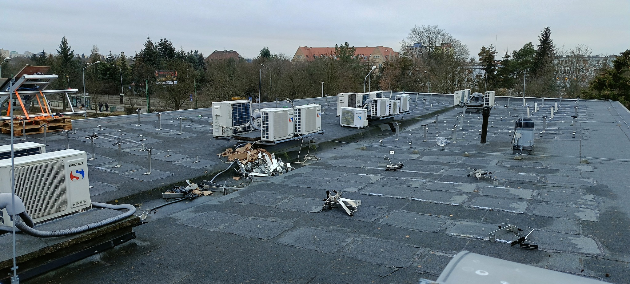 Płaski dach pokryty papą, na którym zamontowano liczne jednostki zewnętrzne klimatyzacji, widoczne pozostałości po pracach montażowych i elementy konstrukcji pod panele słoneczne w tle.