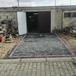 Fragment nowo ułożonej, prostokątnej nawierzchni z ciemnoszarej kostki brukowej z czerwoną obwódką przed garażem z otwartymi drzwiami. W tle sterta kostki i piasku.