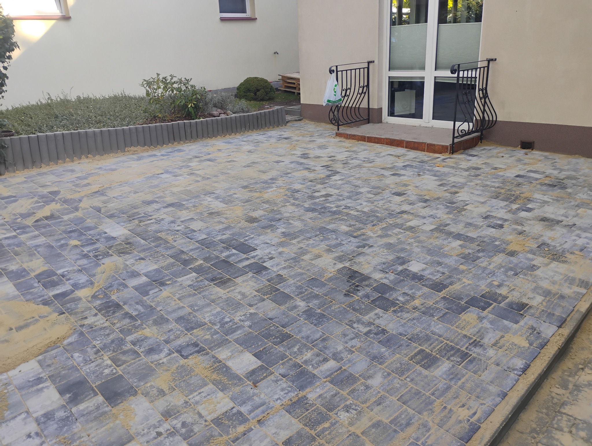 Szara kostka brukowa ułożona na podjeździe, z widocznym piaskiem w szczelinach, obok budynku z metalową balustradą przy wejściu i rabatą z roślinami.