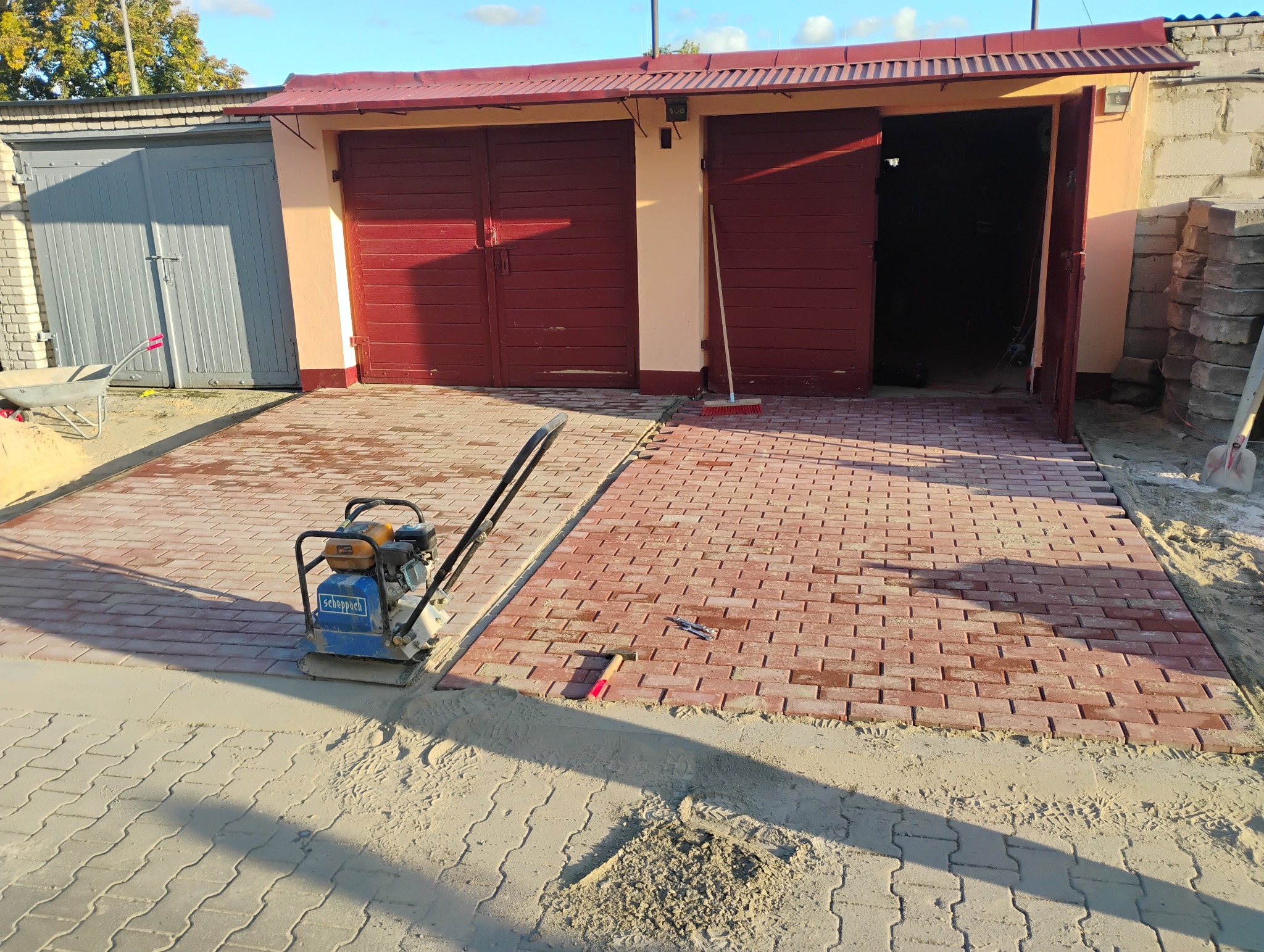 Częściowo ułożona czerwona kostka brukowa przed garażami, widoczna zagęszczarka Scheppach oraz narzędzia brukarskie.