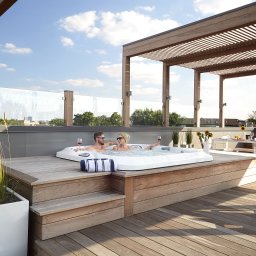 SK Centrum Tarasów - Drewniany taras na dachu z jacuzzi, w którym relaksuje się para z lampkami wina, obok pergola i meble ogrodowe