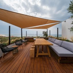 SK Centrum Tarasów - Drewniany taras z widokiem na morze, wyposażony w drewniany stół z ławkami, szarą sofę wbudowaną w ścianę, czarne krzesła i zadaszenie przeciwsłoneczne w kształcie trójkąta.