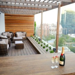 SK Centrum Tarasów - Ekskluzywny taras na dachu z drewnianą pergolą, meblami rattanowymi, drewnianą ścianą, szklaną balustradą, widokiem na miasto i szampanem Moët & Chandon.
