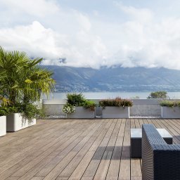 SK Centrum Tarasów - Przestronny taras z drewnianą podłogą, betonowymi donicami z roślinnością, rattanowymi meblami i widokiem na jezioro oraz góry w tle.