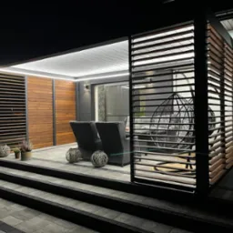 Oświetlona pergola z drewnianymi lamelami i metalową konstrukcją, widoczne meble ogrodowe: dwa fotele i wiszące krzesło, dekoracje roślinne, nocne oświetlenie.