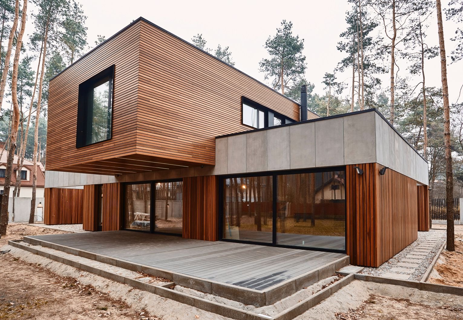 Nowoczesny dom z elewacją z drewnianych paneli, betonowymi elementami i tarasem z desek kompozytowych, otoczony sosnowym lasem.