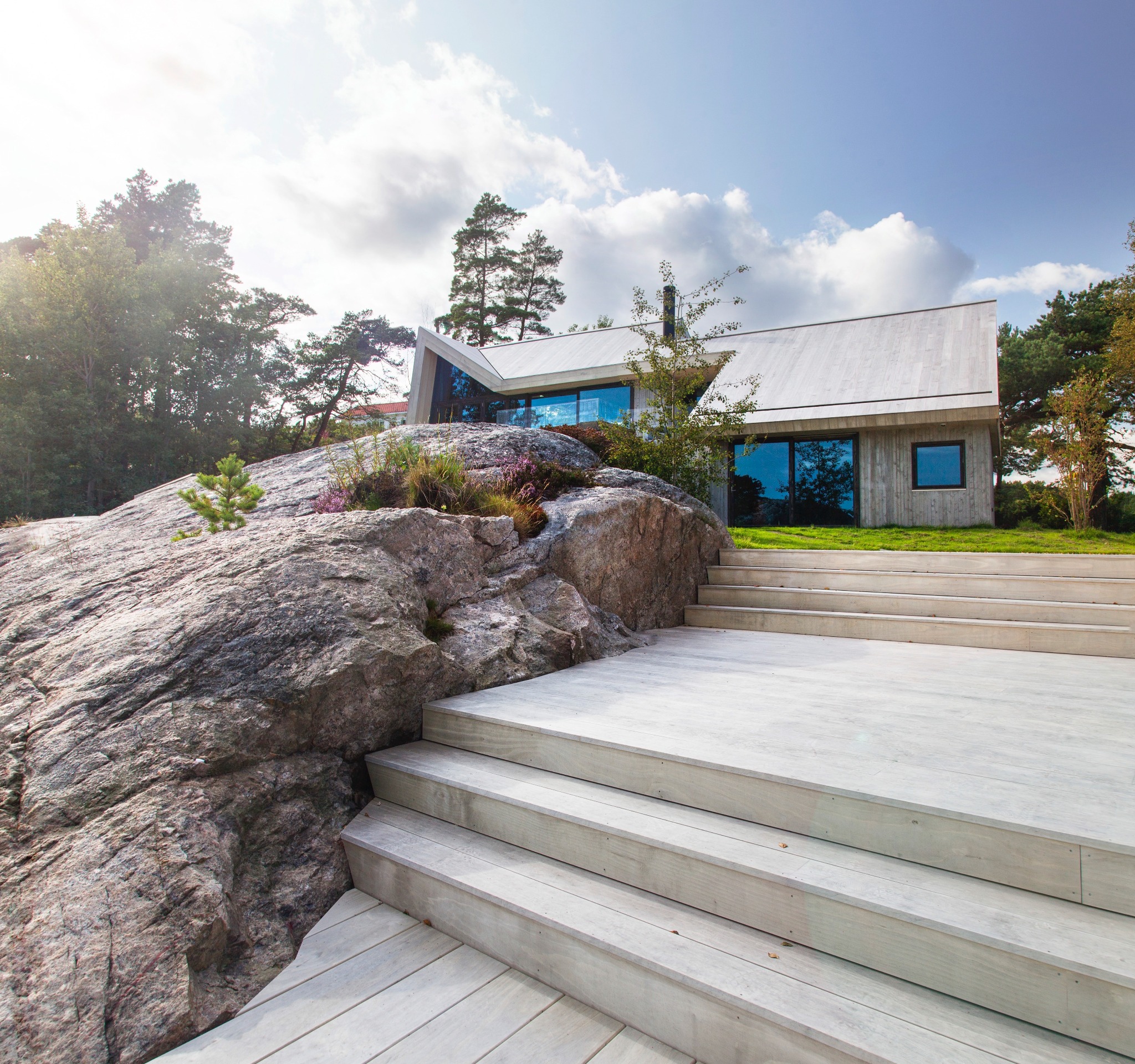 Nowoczesny dom z elewacją z jasnego drewna, zintegrowany z naturalnym krajobrazem, z tarasem z desek i schodami prowadzącymi do budynku, na tle skały porośniętej roślinnością.