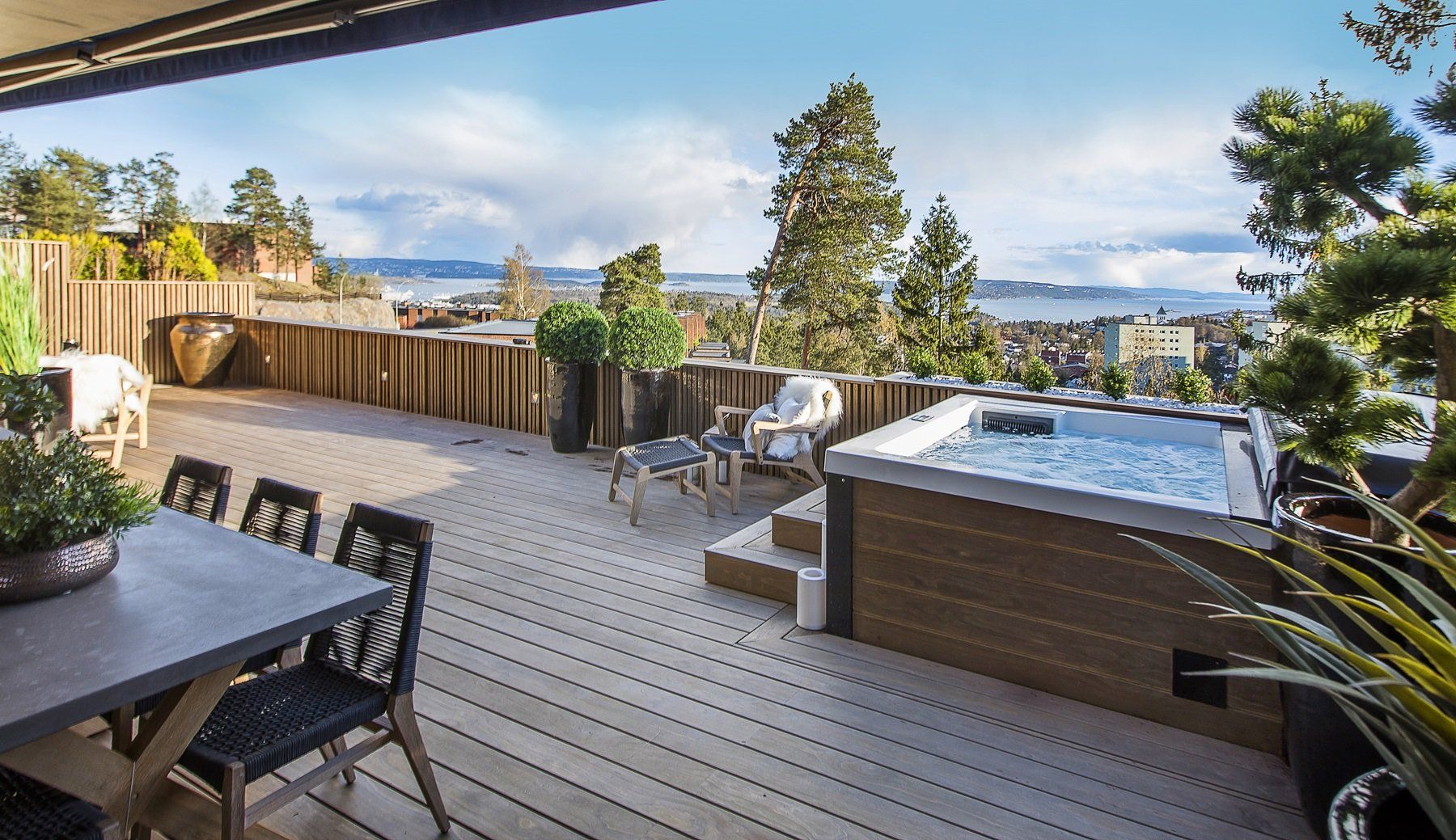 Przestronny taras z drewnianą podłogą, jacuzzi, stołem jadalnym i widokiem na miasto w tle.