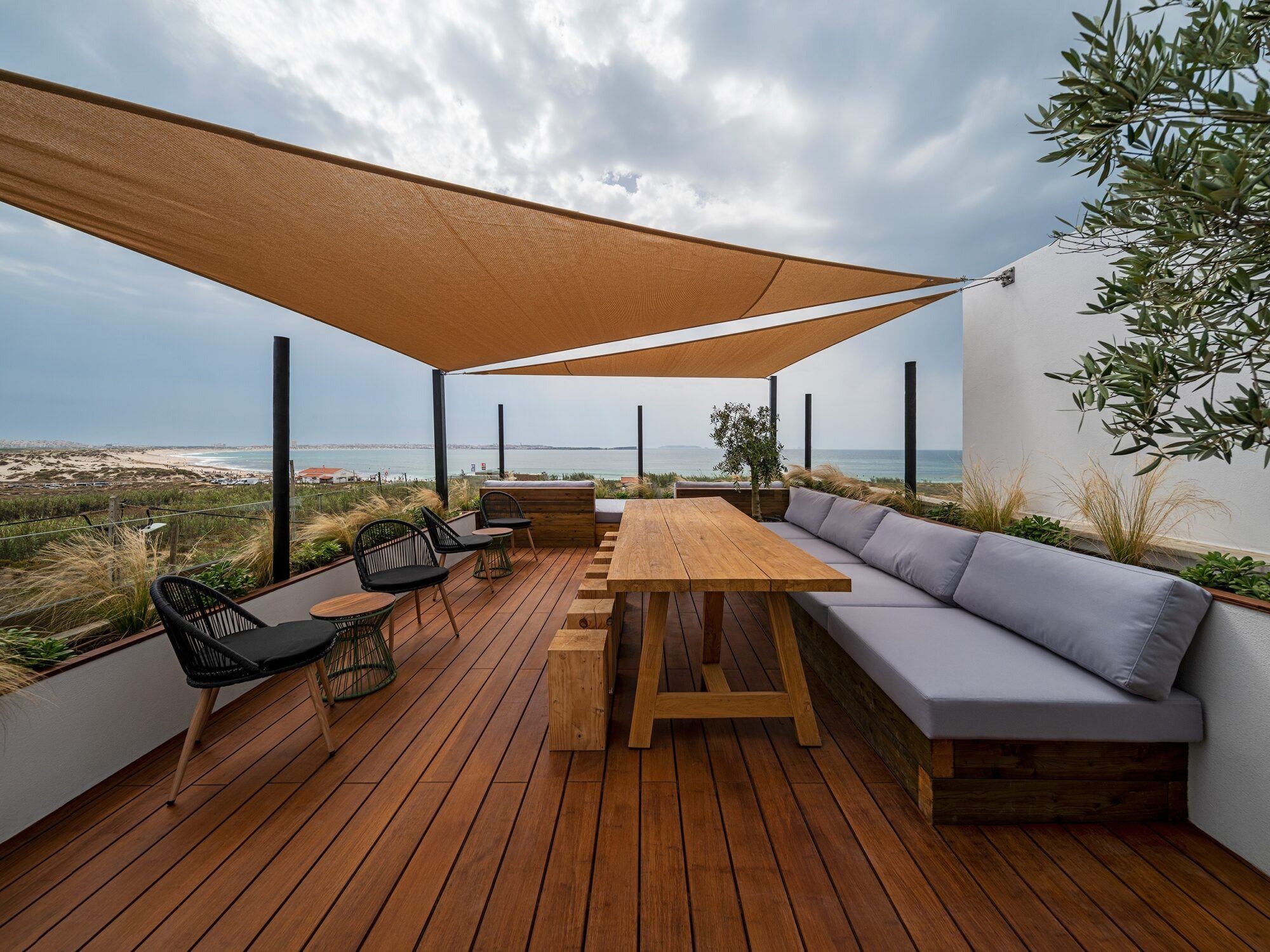 Drewniany taras z widokiem na morze, wyposażony w drewniany stół z ławkami, szarą sofę wbudowaną w ścianę, czarne krzesła i zadaszenie przeciwsłoneczne w kształcie trójkąta.