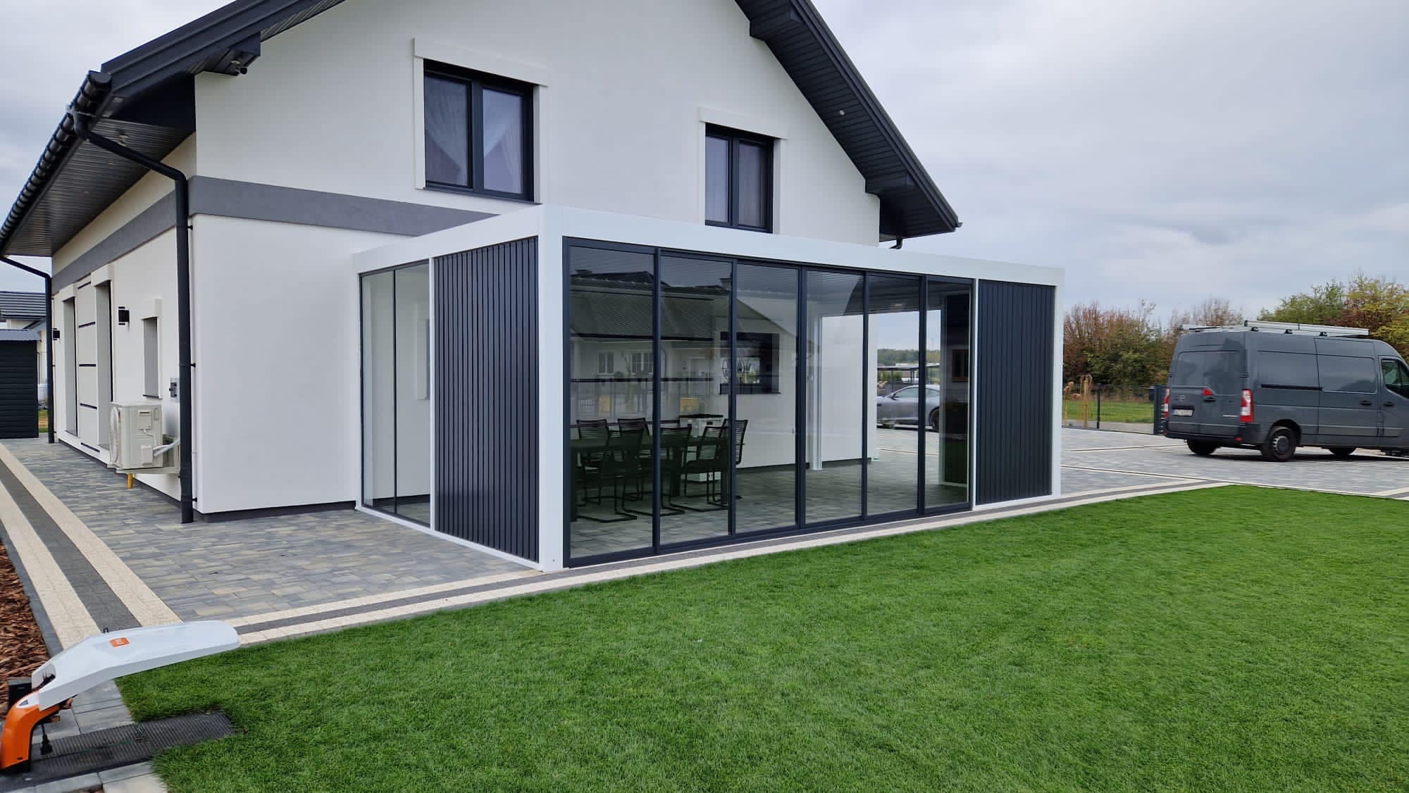 Nowoczesna, przeszklona konstrukcja przylegająca do białego domu z dwuspadowym dachem, z widocznym wnętrzem wyposażonym w stół i krzesła, otoczona trawnikiem i brukowanym podjazdem, z zaparkowanym...