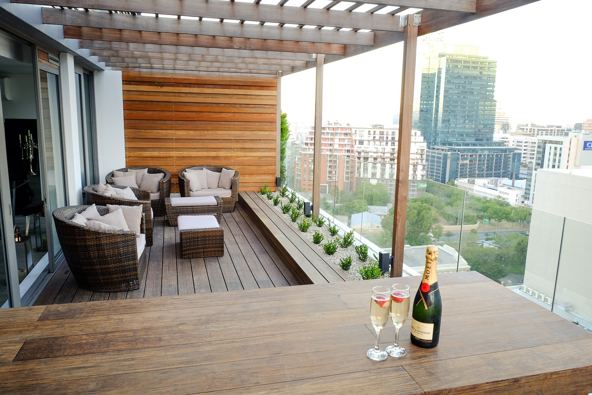 Ekskluzywny taras na dachu z drewnianą pergolą, meblami rattanowymi, drewnianą ścianą, szklaną balustradą, widokiem na miasto i szampanem Moët & Chandon.