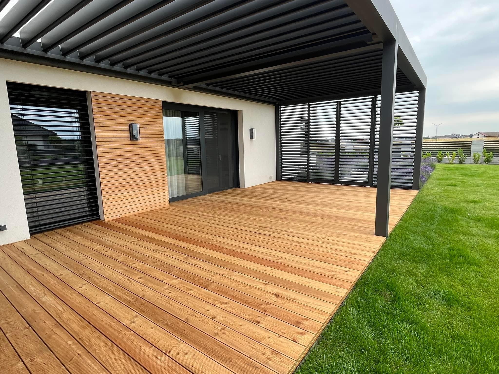 Nowoczesny taras z drewna z czarną metalową pergolą, przylegający do domu z drewnianą elewacją i dużymi oknami, otoczony zadbanym trawnikiem.